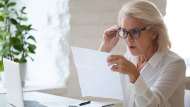 Surprised aged woman worker in glasses sit at office desk reading paper document or contract feel confused with bad news, frustrated senior businesswoman stunned by received paperwork correspondence