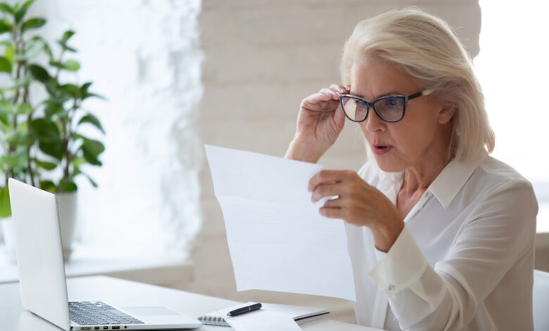 Surprised aged woman worker in glasses sit at office desk reading paper document or contract feel confused with bad news, frustrated senior businesswoman stunned by received paperwork correspondence