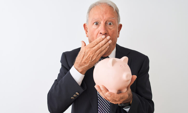 Senior grey-haired businessman holding piggy bank standing over isolated white background cover mouth with hand shocked with shame for mistake, expression of fear, scared in silence, secret concept
