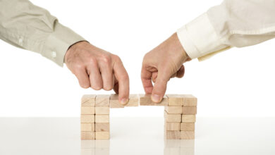 Conceptual image of business merger and cooperation - two male hands joining effort to build a bridge of wooden pegs on a white desk with reflection over white background.
