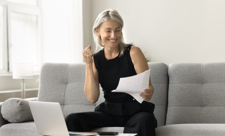 Happy successful mature investor woman celebrating profit, income from investment, retirement savings, reading financial paper document, making winner hand, smiling, sitting at laptop at home