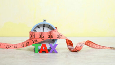 Letters on Tax standing against the alarm clock wrapped with measuring tape on the wooden table.